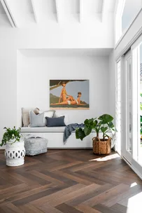 Cozy white corner with cushions and painting, herringbone wood floor, large leafy plant, and natural light from windows.