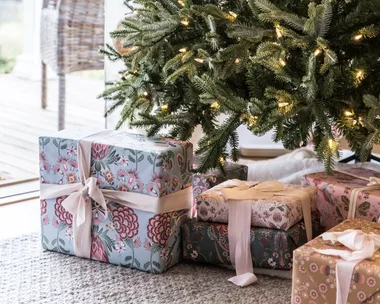 Christmas tree with lights and wrapped gifts in floral paper underneath on a carpeted floor near a window.