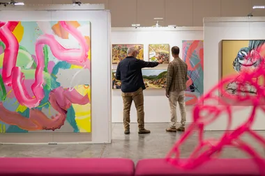 Two people view colorful abstract and landscape paintings in an art gallery, with a pink sculpture in the foreground.