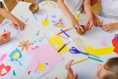 Children's hands crafting with paper, markers, and scissors on a white table.