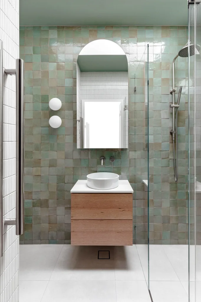 An ensuite bathroom with green zellige handmade wall tiles, two round wall sconces, a curved basin, tapware in brushed nickel, wooden joinery and white kitkat wall tiles. A shower is beside the vanity.