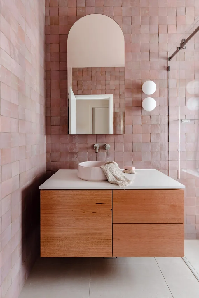 A main bathroom featuring pink zellige wall tiles, white floor tiles, wooden joinery, a round basin, an arched mirror and two round wall sconces beside a shower. The tapware is brushed nickel in a curved style. The bathroom design is cohesive.