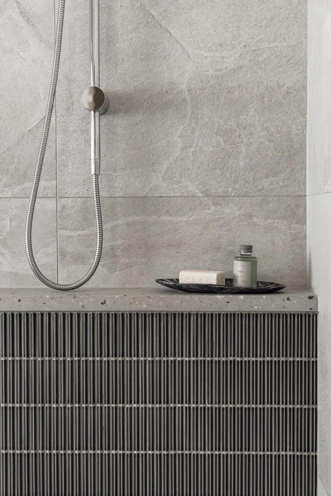 A close up shot of details in a shower, complete with grey stone wall tiles. The same material is used to create a ledge, which products have been placed upon. Beneath this are grey kitkat tiles while the shower tapware is brushed nickel.