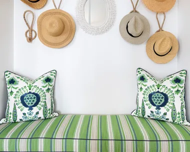 A built in bench with a white base, striped green cushion and woven hats displayed on the wall as decor.