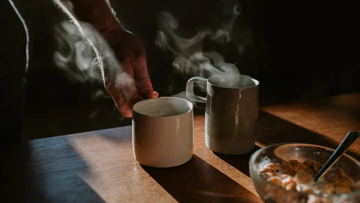 Two cups of coffee on a wooden table with a bowl of breakfast cereal. Someone lifts one of the mugs out of frame.