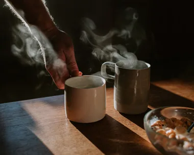 Two cups of coffee on a wooden table with a bowl of breakfast cereal. Someone lifts one of the mugs out of frame.