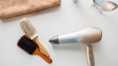 Hairdryer, round brush, paddle brush, towel, and mirror on a white surface.