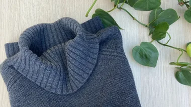 Folded gray turtleneck sweater on a light wooden surface next to green vines.