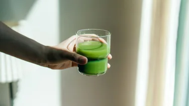 Hand holding a glass of green juice with soft light in the background.