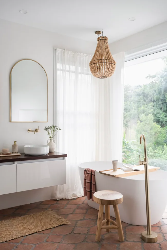 A mediterranean coastal style bathroom with a large white freestanding bath, brass hardware, arched mirror and view on to surrounding bushland.