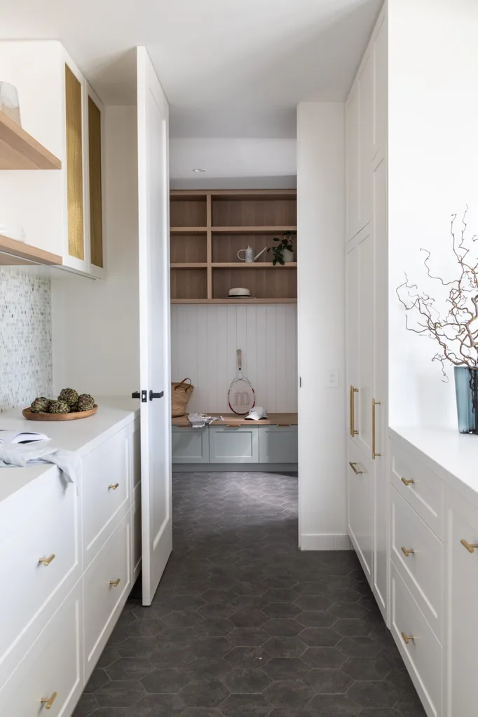 A butler's pantry with dark tiled flooring, white joinery on either side of the galley layout and brass mesh profiles on the upper cabinetry. The butler's pantry leads to a mud room with timber joinery.