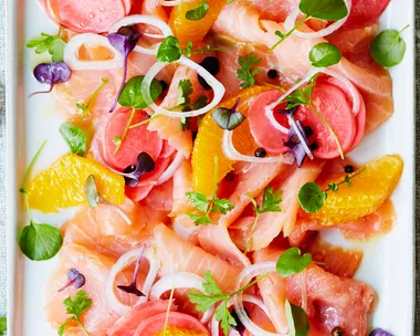 Plate of cured salmon with citrus, radish, microgreens, and onion rings, next to a stack of small forks on a linen cloth.