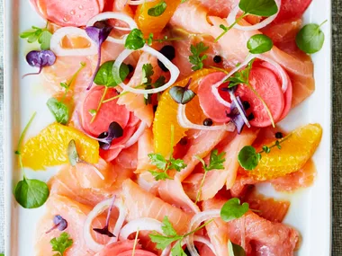Plate of cured salmon with citrus, radish, microgreens, and onion rings, next to a stack of small forks on a linen cloth.